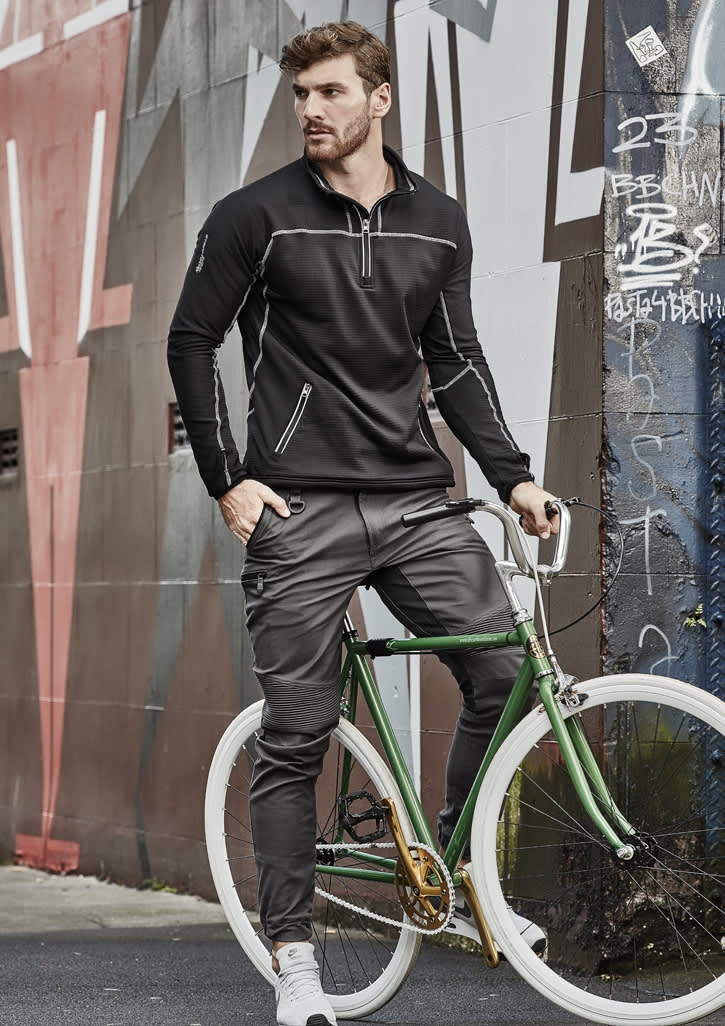 A man in black pullover and grey cuffed stretch pants poses with a green bicycle against a graffiti wall.