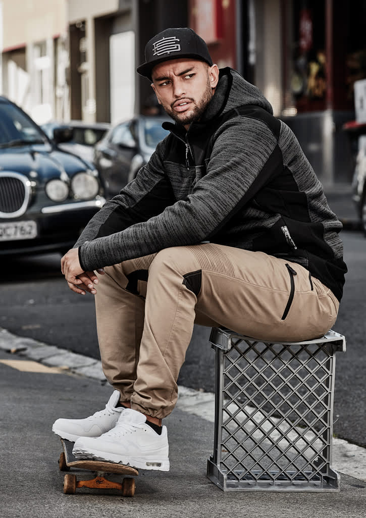 A man sits on a crate wearing cuffed beige stretch pants and a black hoodie. He has white sneakers and a cap.