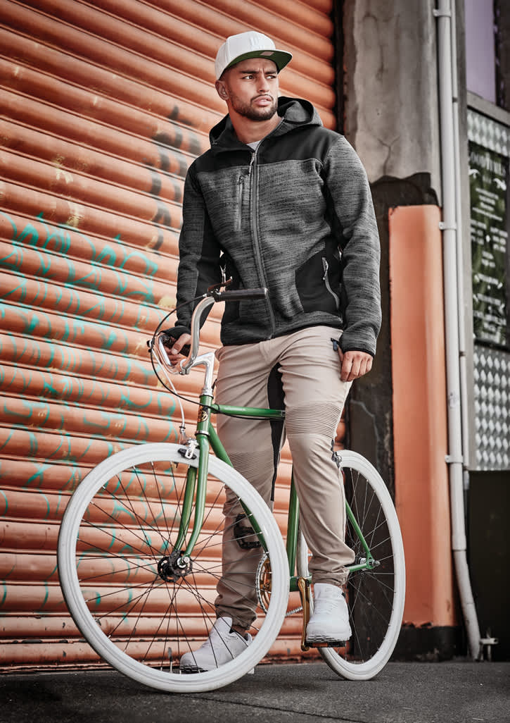 A man wearing cuffed beige stretch pants and a grey jacket stands beside a green bicycle against a textured orange backdrop.