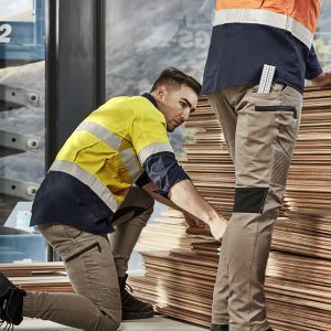 Two workers in high-visibility jackets and stretch cuffed pants, stacked wood planks in an industrial setting.