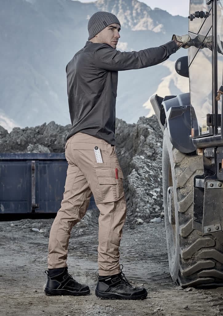 Men's curved cargo pants in beige, featuring multiple pockets, paired with dark boots and a grey beanie against a rugged backdrop.