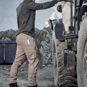 A man wearing beige curved cargo pants and a long-sleeve shirt stands next to a truck in a rugged outdoor setting.