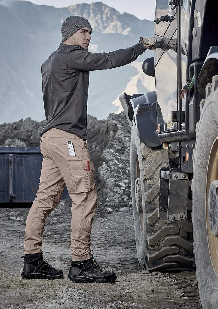A man wearing beige curved cargo pants and a long-sleeve shirt stands next to a truck in a rugged outdoor setting.