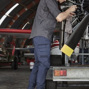 Mens Streetworx Heritage Pant in navy, paired with tan work boots, worn by a man repairing machinery in a hangar.