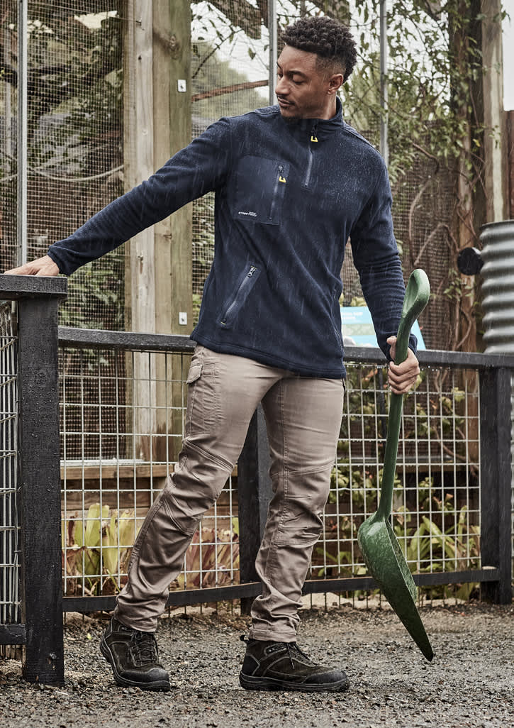 A man wearing beige work pants and a navy fleece jacket, holding a green gardening tool in an outdoor setting.