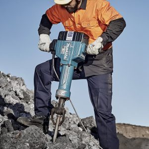 A man in an orange and black jacket and ear protection operates a blue jackhammer on rocky terrain.