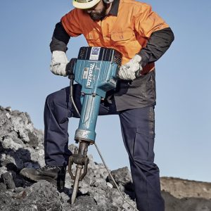 A man in safety gear operates a jackhammer on a rocky surface, wearing an orange shirt and navy blue cargo pants.