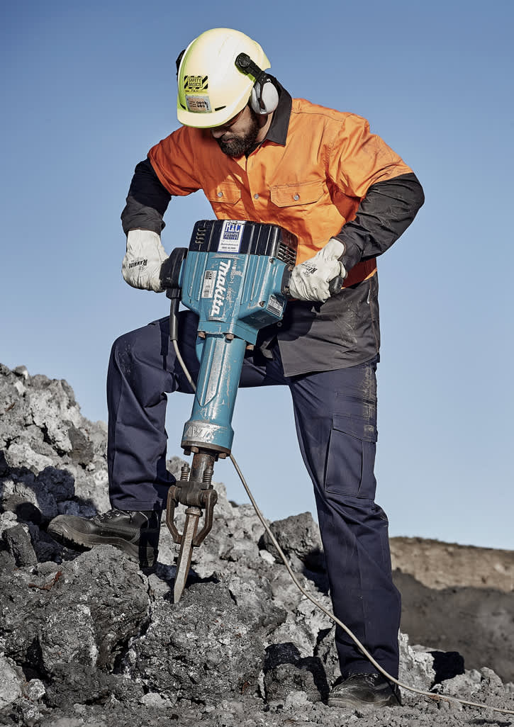 A man in safety gear operates a jackhammer on a rocky surface, wearing an orange shirt and navy blue cargo pants.