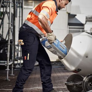 A man in orange and grey high-visibility workwear pours from a bag into a mixer, wearing navy rugged cargo pants.