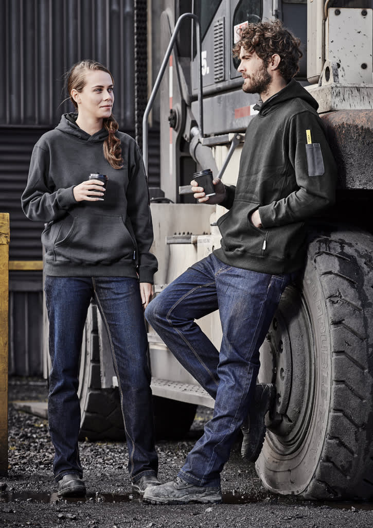 A man and woman wear black hoodies and blue denim work jeans, standing beside a large vehicle in an industrial setting.