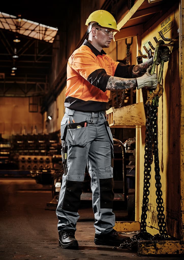 A man in an orange and grey shirt and multi-pocket grey pants works in a warehouse, wearing safety gear and a helmet.