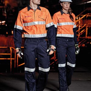 Two individuals wearing high-visibility orange shirts and navy cargo pants walk in an industrial setting, both wearing helmets and gloves.