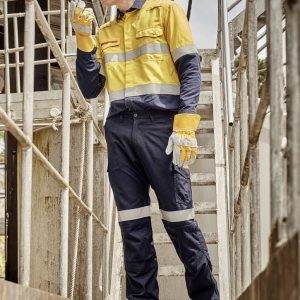 A worker in a yellow and navy work shirt and cargo pants, wearing gloves and a hard hat, on a metal staircase.