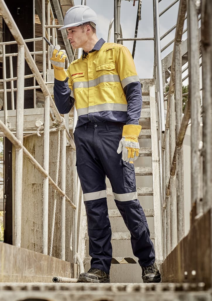 A worker in a yellow and navy work shirt and cargo pants, wearing gloves and a hard hat, on a metal staircase.