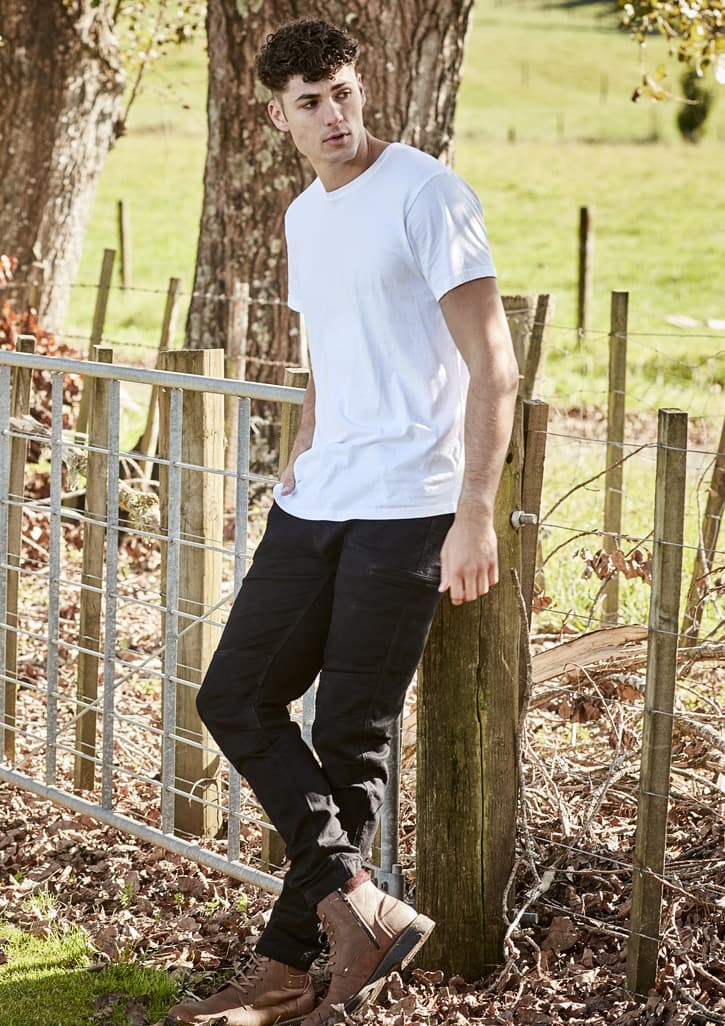 A person wearing black stretch jeans and a white t-shirt stands by a wooden fence in a grassy area.