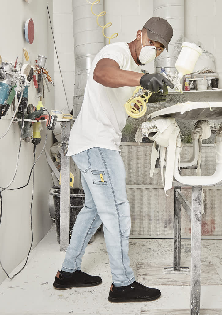A man wearing light blue stretch jeans and a white t-shirt is spray painting in a workshop.