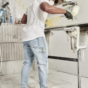 A man wearing a white shirt and grey streetwear stretch jeans, painting in a workshop setting.