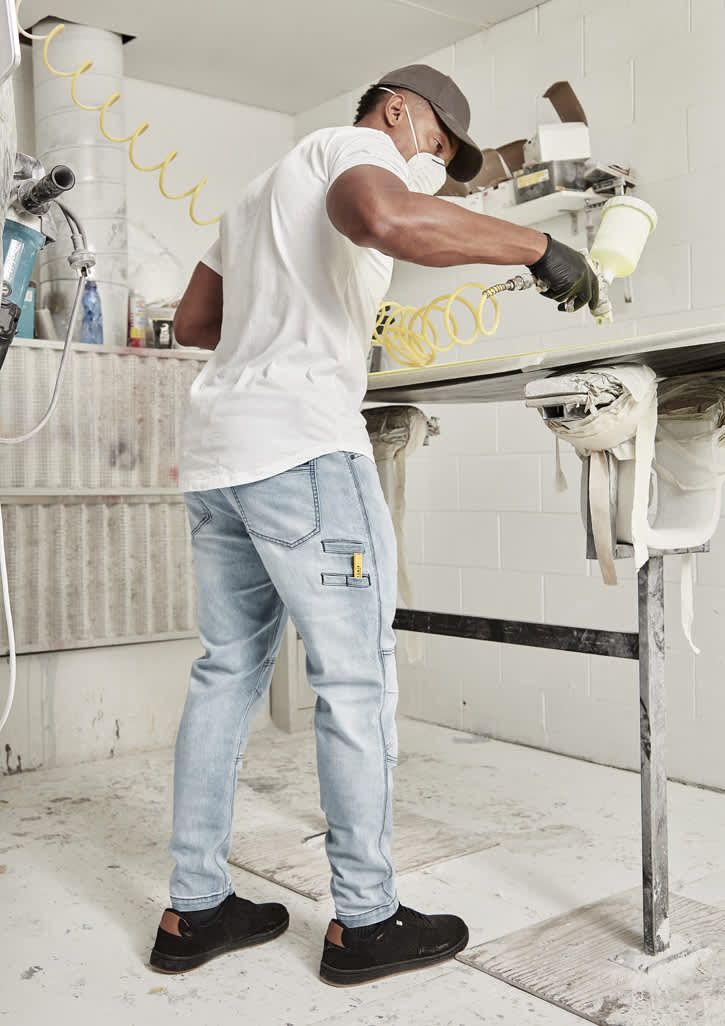 A man wearing a white shirt and grey streetwear stretch jeans, painting in a workshop setting.