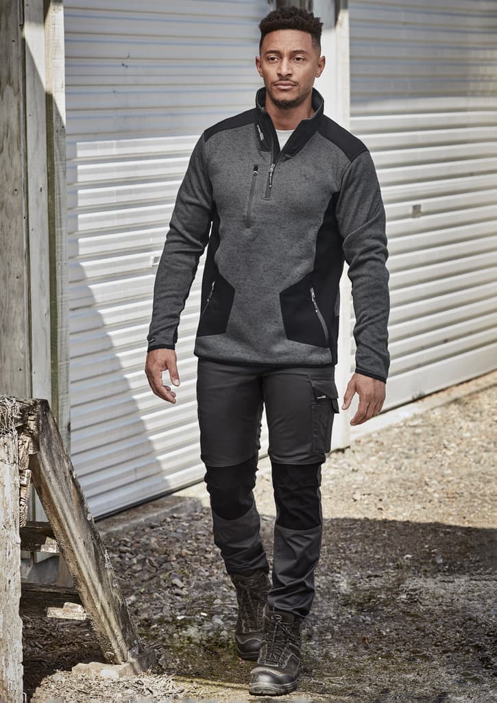 A man in dark grey and black workwear pants and a fitted grey pullover, walking in an outdoor setting.
