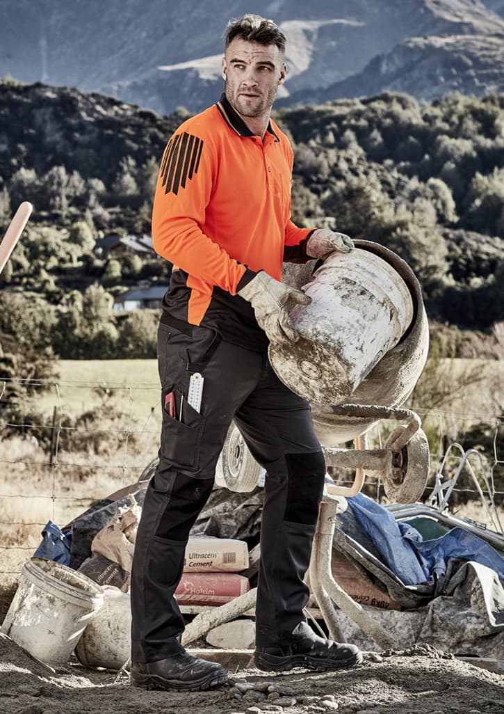 A man in an orange shirt and black tough work pants carries a bucket at a construction site, with mountains in the background.