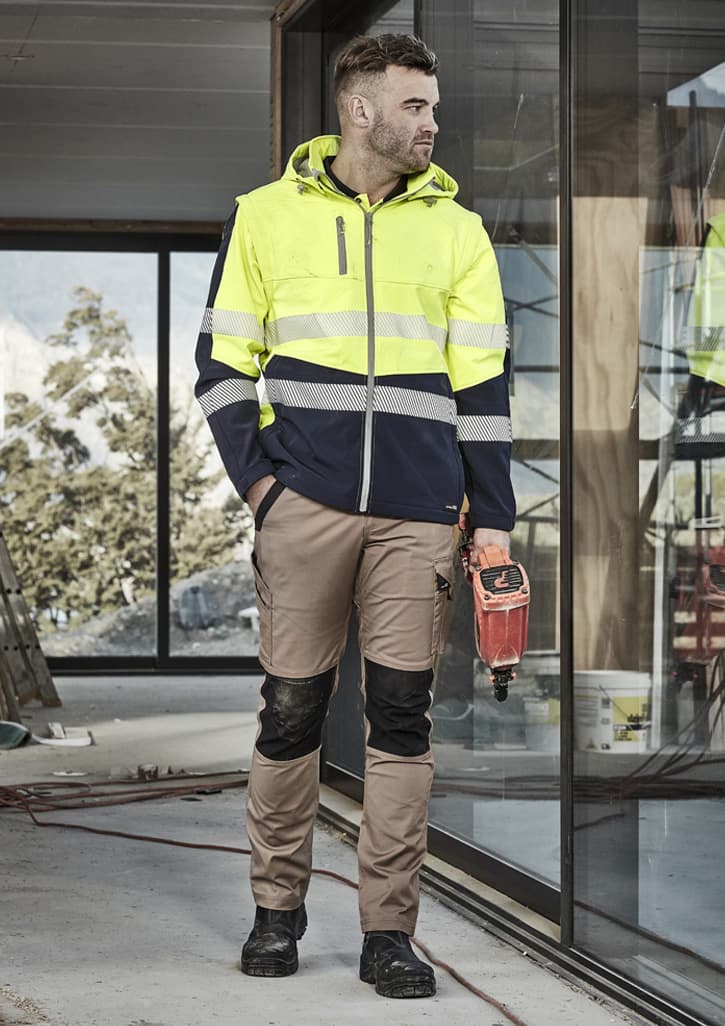 A man wearing a high-visibility yellow and navy work jacket and khaki work pants, standing in a construction setting.
