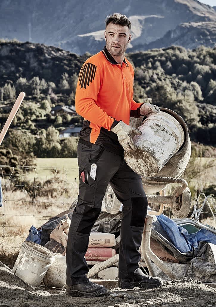 A worker in an orange high-visibility shirt and black tough pants, holding a bucket on a job site with mountains in the background.