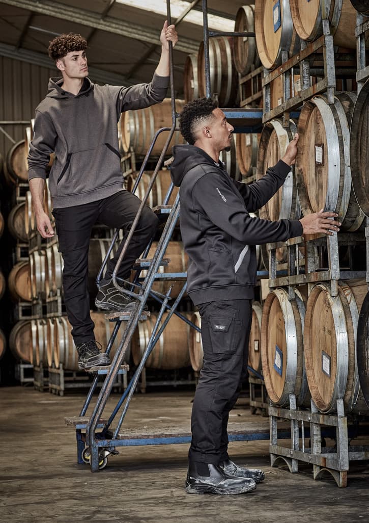 Two men in rugged cooling stretch pants, one standing on a ladder and the other inspecting wooden barrels, in a warehouse setting.