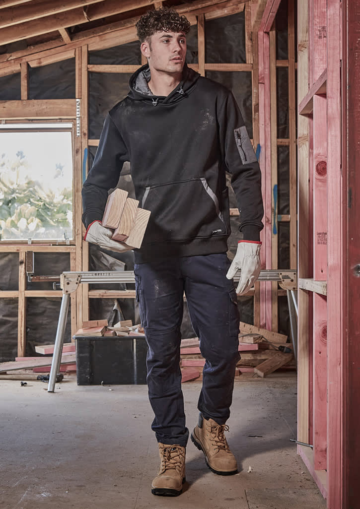 A man wearing black cargo pants and a black hoodie is holding wooden blocks, standing in a construction setting.