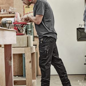 Men's rugged cooling stretch pants in black, featuring cargo pockets, are worn by a person working in a workshop.