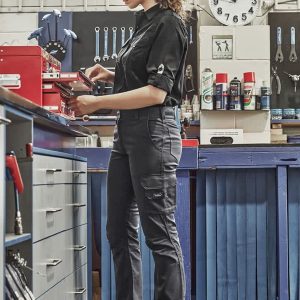 A woman in black rugged cargo pants stands at a workshop bench, engaging with tools and equipment. The pants feature pockets and a logo.