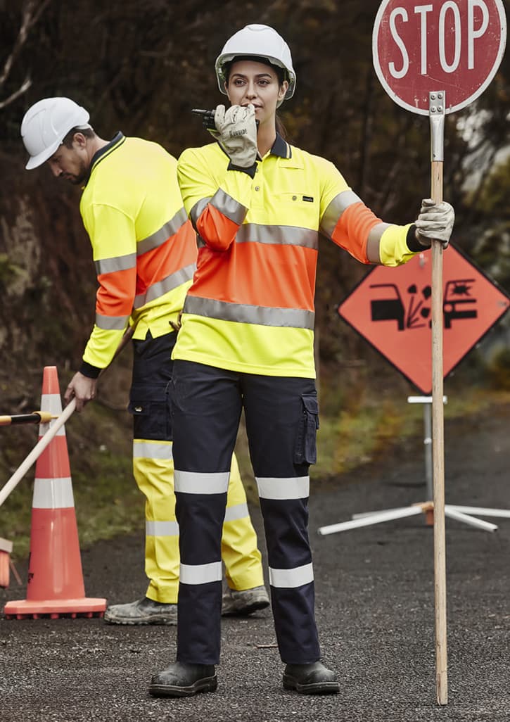 A woman in a high-visibility yellow and orange work shirt and dark pants holds a stop sign. A man in the background wears similar attire.