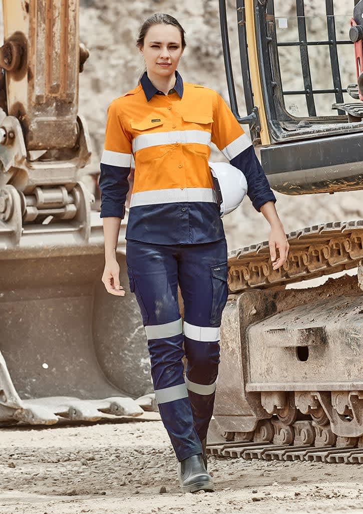 A woman in an orange and navy work outfit with reflective stripes stands near heavy machinery. The outfit includes cargo pants.