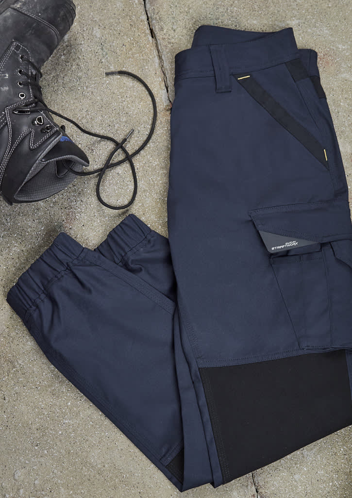 A pair of navy women's work pants laid flat, featuring pockets and an elastic waistband, beside a work boot.
