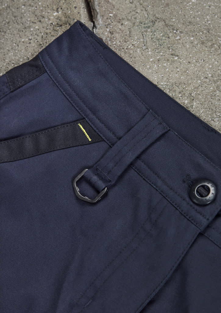 A close-up of navy work pants featuring a belt loop, button closure, and a side black panel. Contains a branded logo.