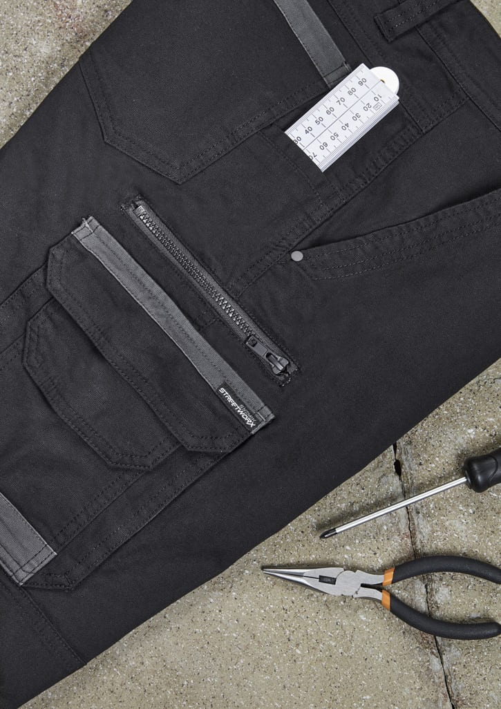 A pair of black work pants featuring multiple pockets, a zipper, and a branding logo. Tools are placed beside them.
