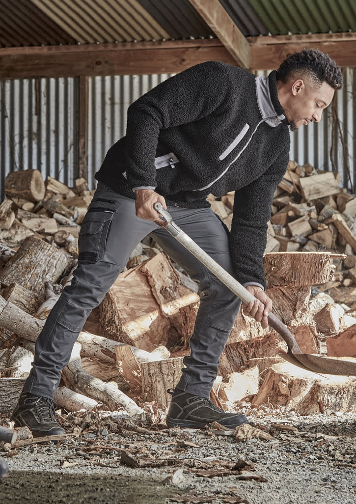 A model is wearing black work pants and a black fleece top while chopping wood in a rustic setting.