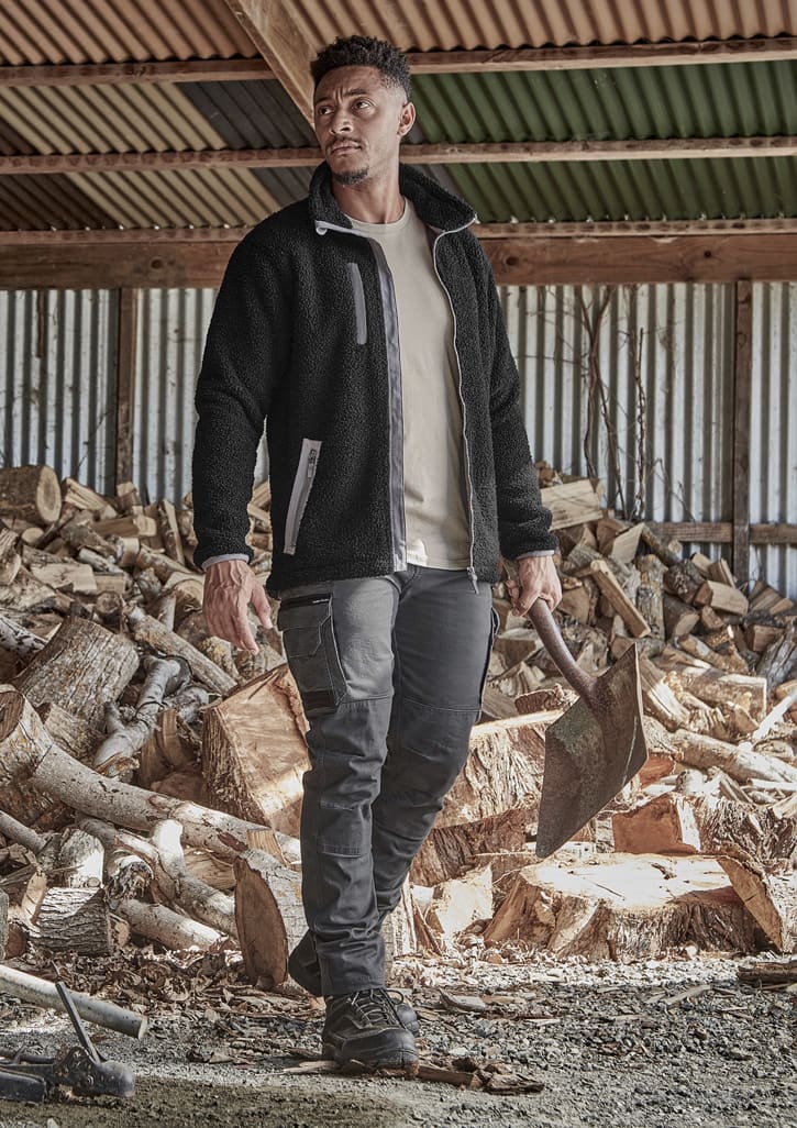 A model wearing black work pants and a black fleece jacket stands outdoors, with stacks of timber in the background.