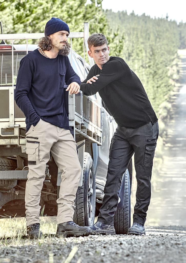Two men wearing work pants in beige and black, standing beside a vehicle in a forested area. Both have casual attire and a logo on the clothing.