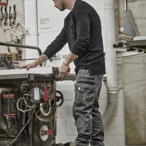 A man wearing black work pants stands by a workshop table, handling tools in a well-equipped space.