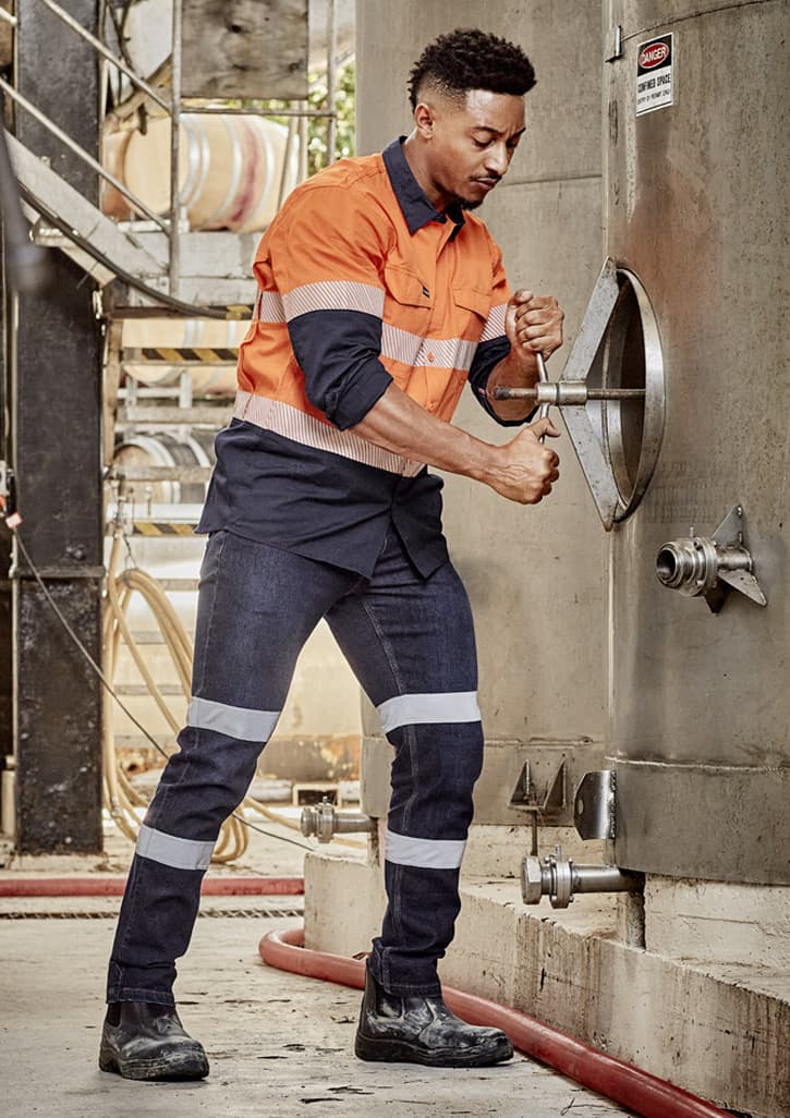 A man in an orange and navy work shirt and blue stretch jeans adjusts equipment, featuring reflective stripes and safety boots.