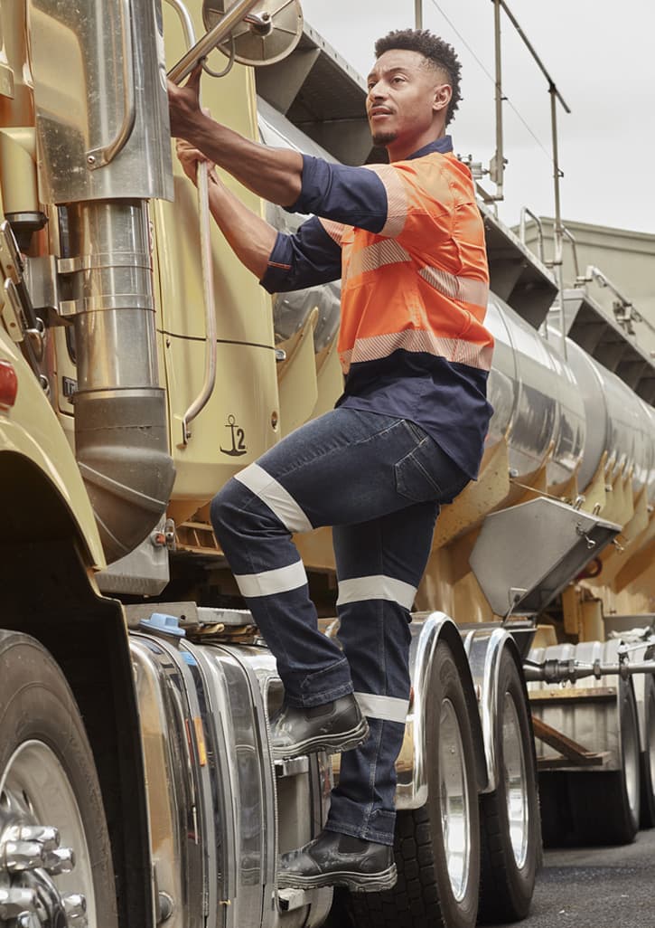 A man in an orange and navy work shirt climbs onto a truck, wearing dark blue stretch jeans with reflective stripes.