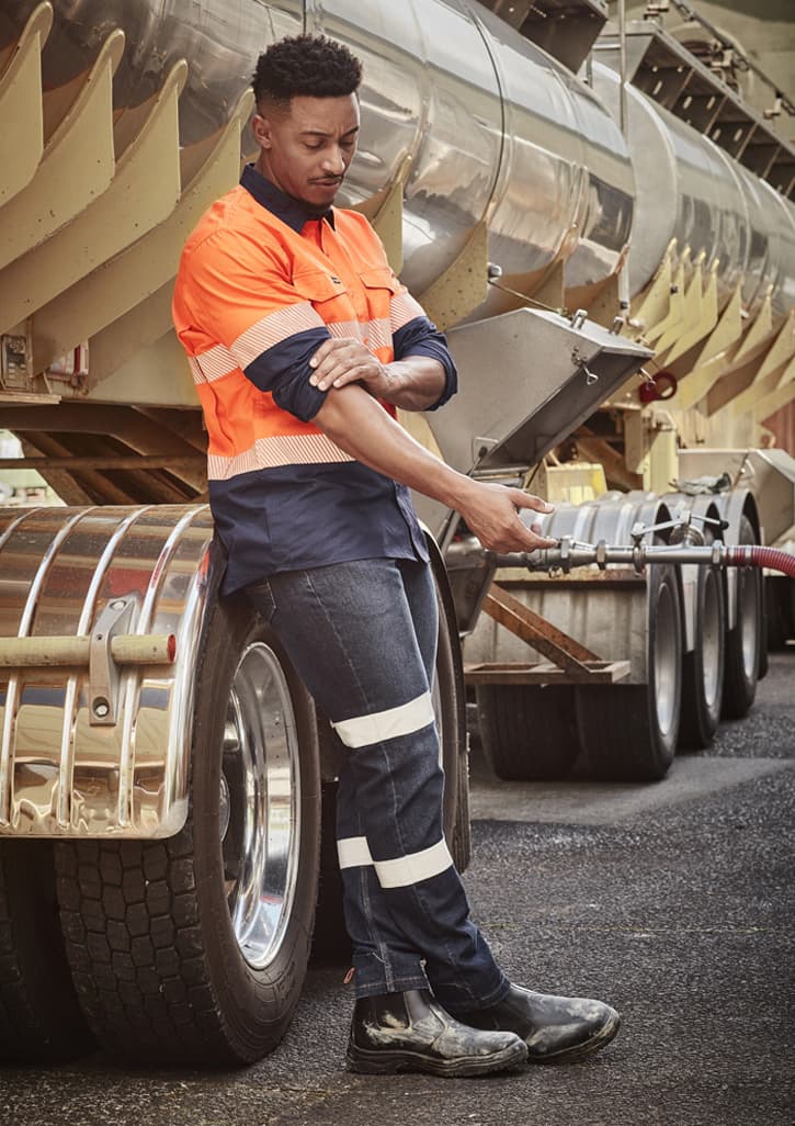 A man in a high-visibility orange and navy work shirt and dark stretch jeans leans against a truck.