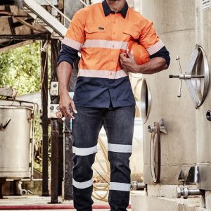A man in orange and navy workwear with reflective stripes, holding a hard hat, stands in an industrial setting.