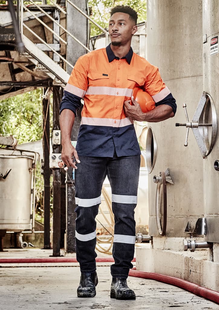 A man in orange and navy workwear with reflective stripes, holding a hard hat, stands in an industrial setting.