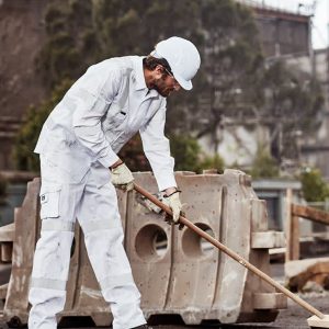 A man in white work pants and a helmet is using a broom on a construction site. The pants have a branding logo.