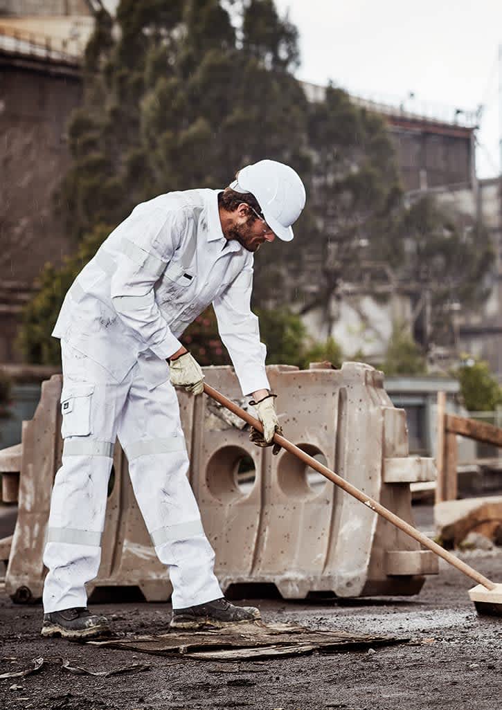 A man in white work pants and a helmet is using a broom on a construction site. The pants have a branding logo.