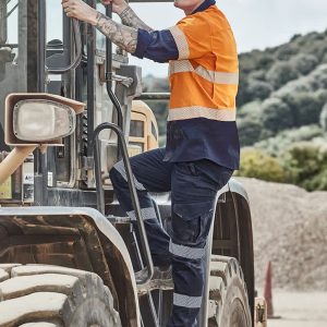 A man wearing an orange and navy shirt and dark work pants climbs into a large machinery vehicle on a worksite.