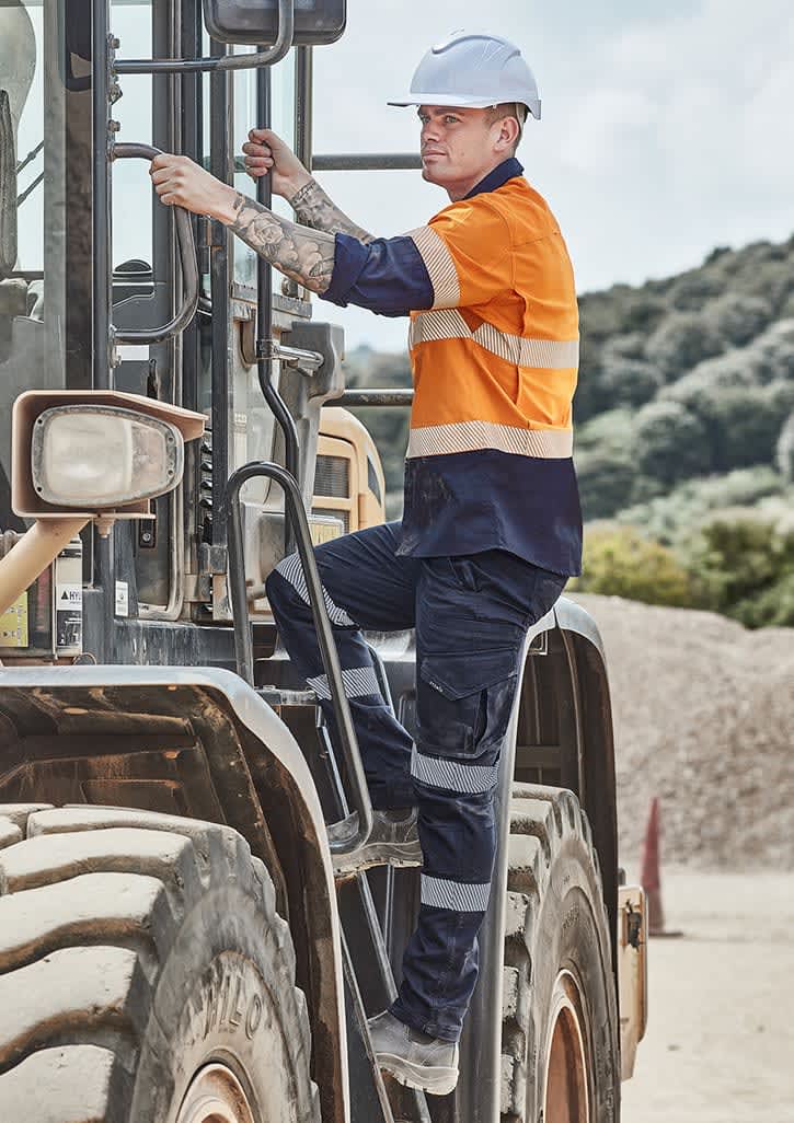 A man wearing an orange and navy shirt and dark work pants climbs into a large machinery vehicle on a worksite.