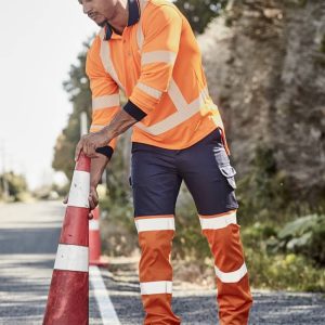 A man in high-visibility orange and navy work pants adjusts a traffic cone on a road. The pants feature reflective strips.