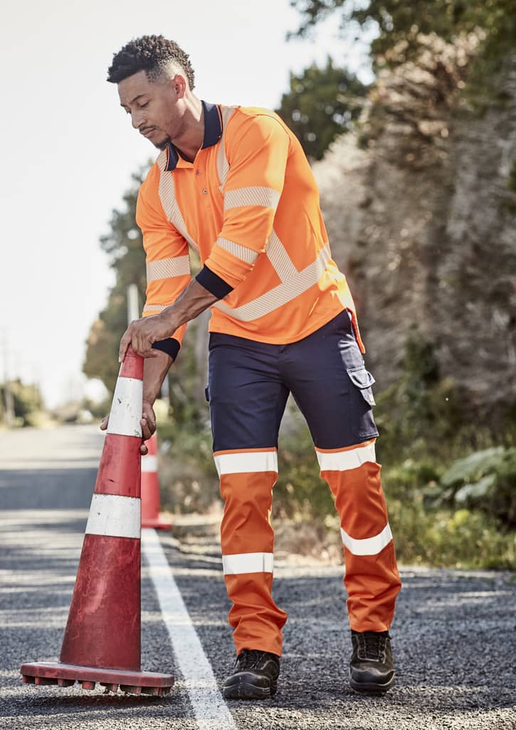 A man in high-visibility orange and navy work pants adjusts a traffic cone on a road. The pants feature reflective strips.